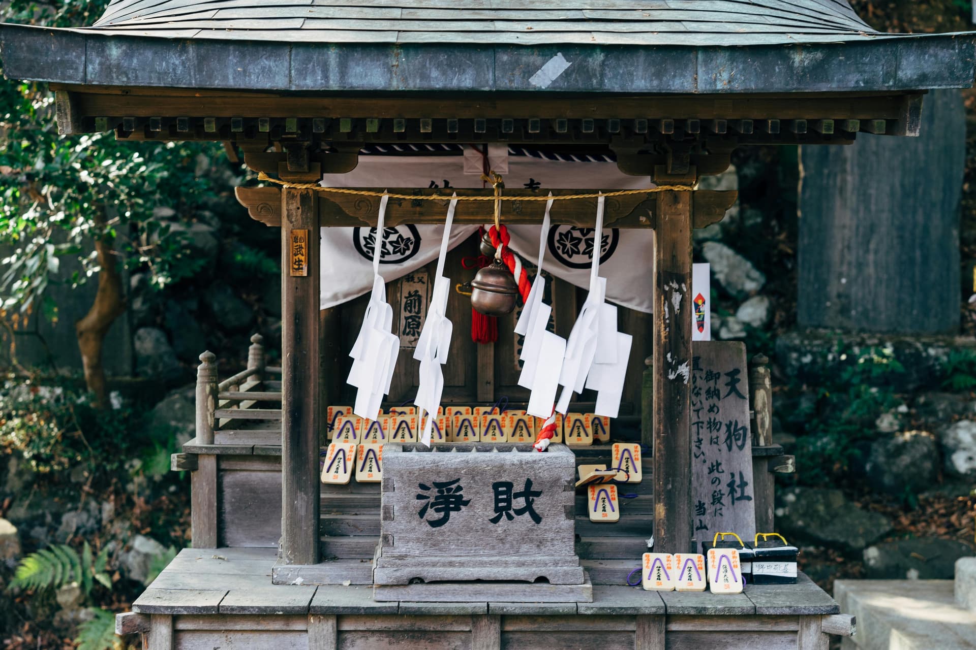 Traditional Japanese shrine with ema prayer tablets, shide paper streamers, and purification bell surrounded by lush greenery