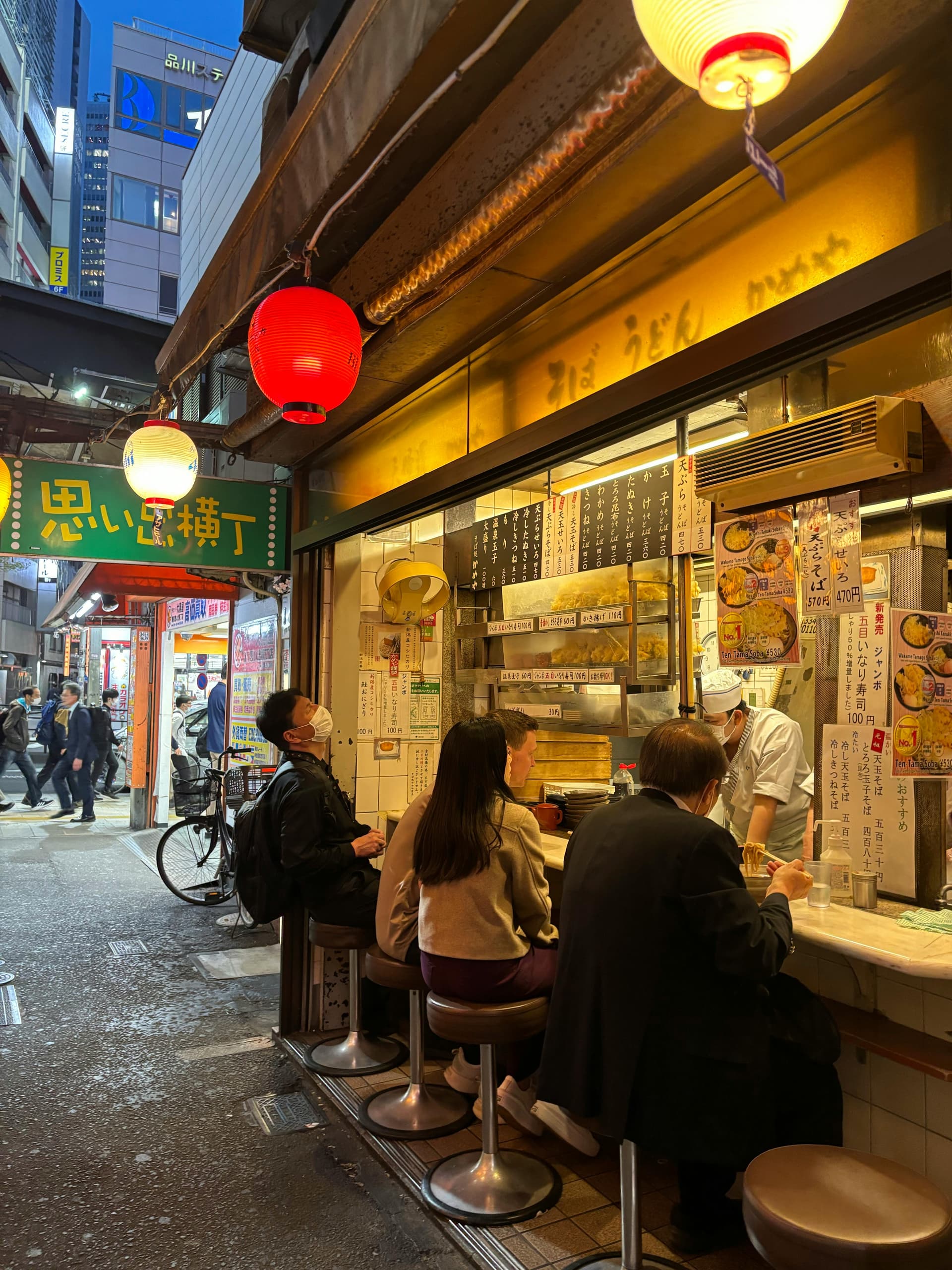 Patrons seated at an outdoor counter stall under red lanterns in Omoide Yokocho
