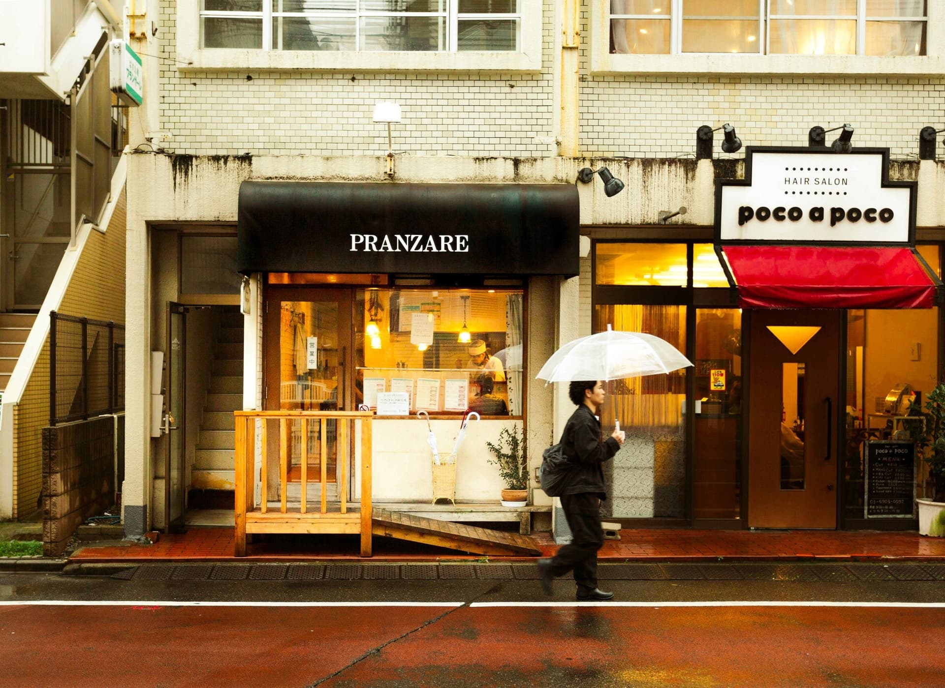 Solo figure with umbrella walking past a warmly lit restaurant on a rainy side street