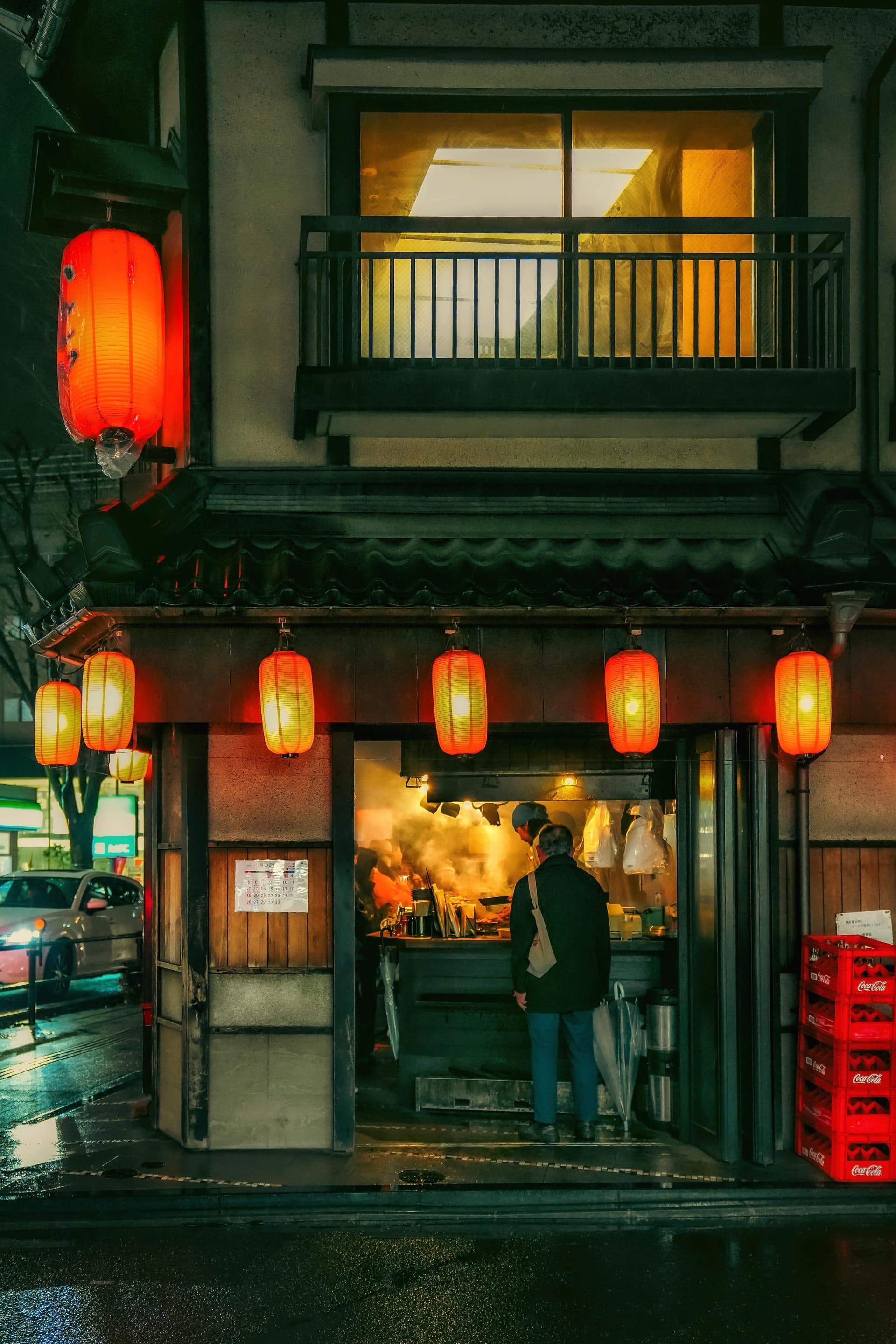 Solo figure ordering at a smoky yakitori stall under red lanterns in the rain