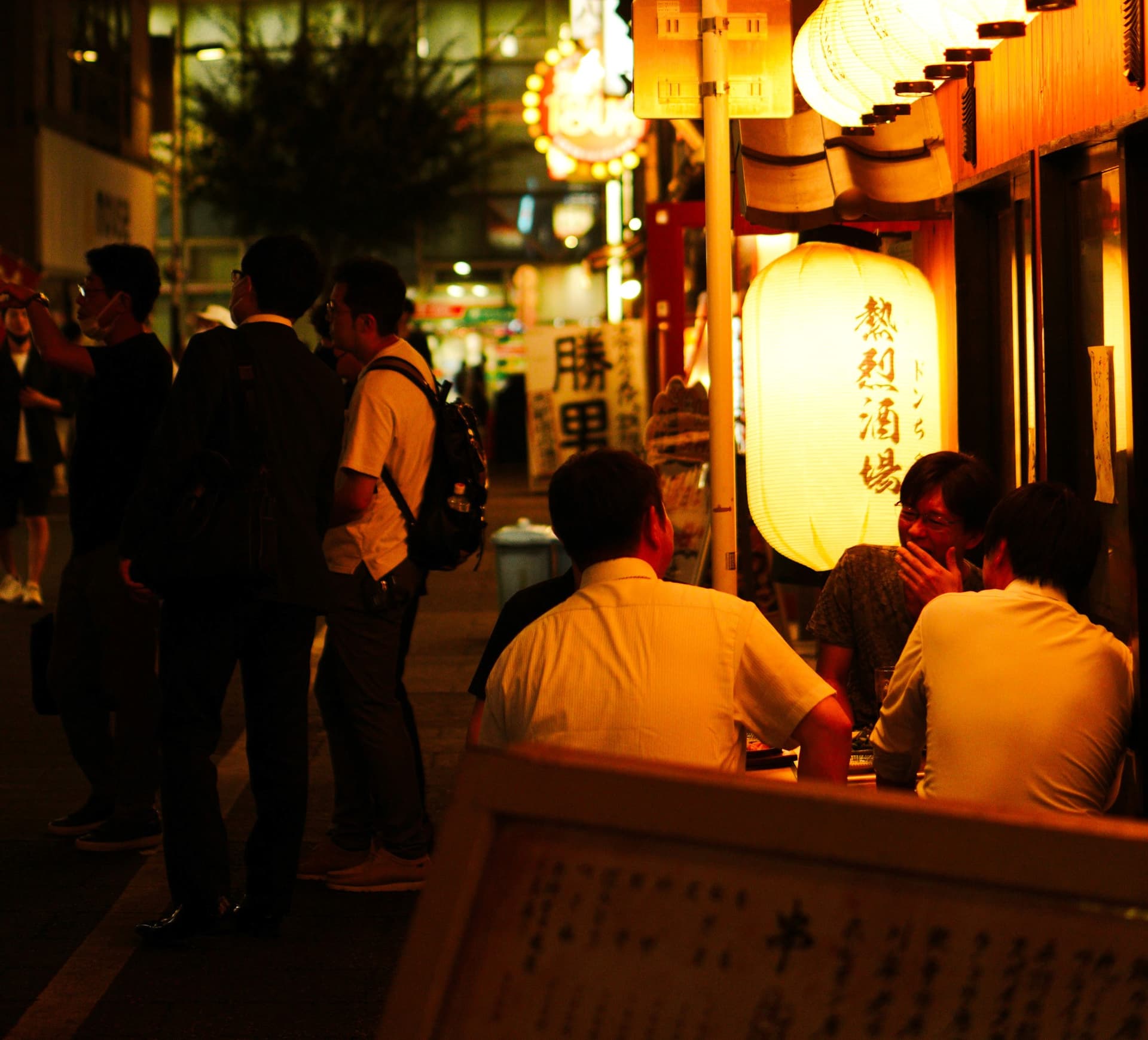 Groups of salarymen outside a lantern-lit izakaya strip in the evening