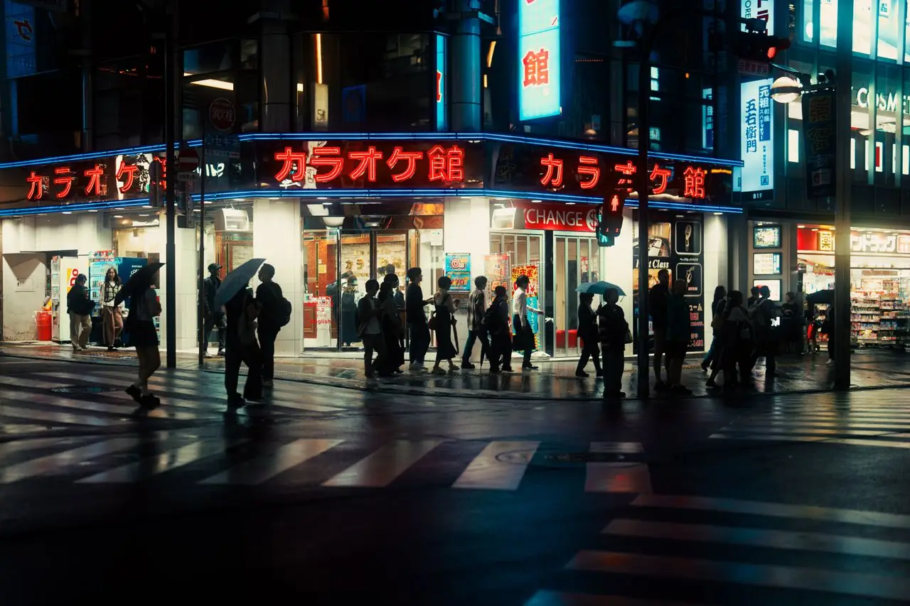 Pedestrians with umbrellas crossing in front of a Karaoke Kan building on a rainy Tokyo night