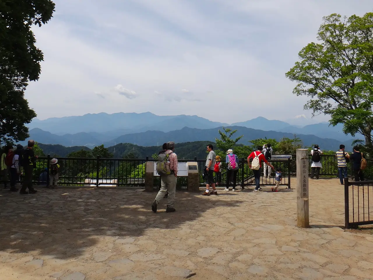 Hikers at the Mt Takao summit viewpoint with hazy mountains and a faint Fuji beyond