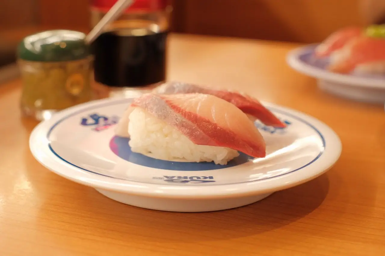 Two pieces of nigiri sushi on a branded Kura Sushi plate beside soy sauce