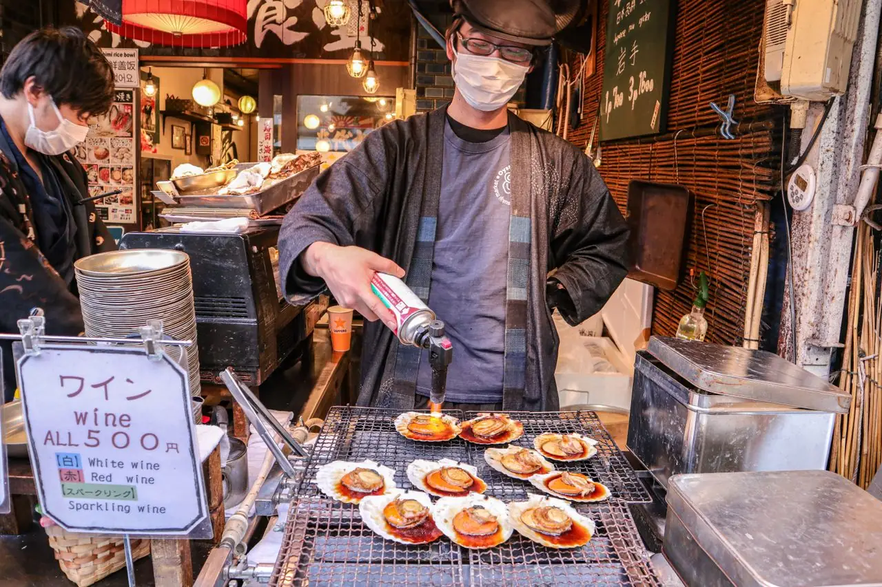 A vendor torch-grills scallops in their shells at a market food stall