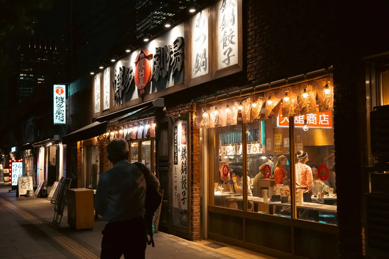 A passerby walking past a brightly lit restaurant front on a Tokyo street at night