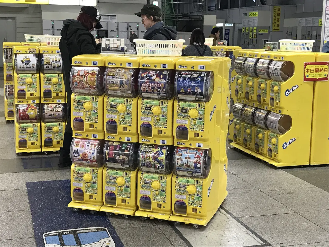 Rows of yellow gachapon capsule toy machines on a Japanese station concourse