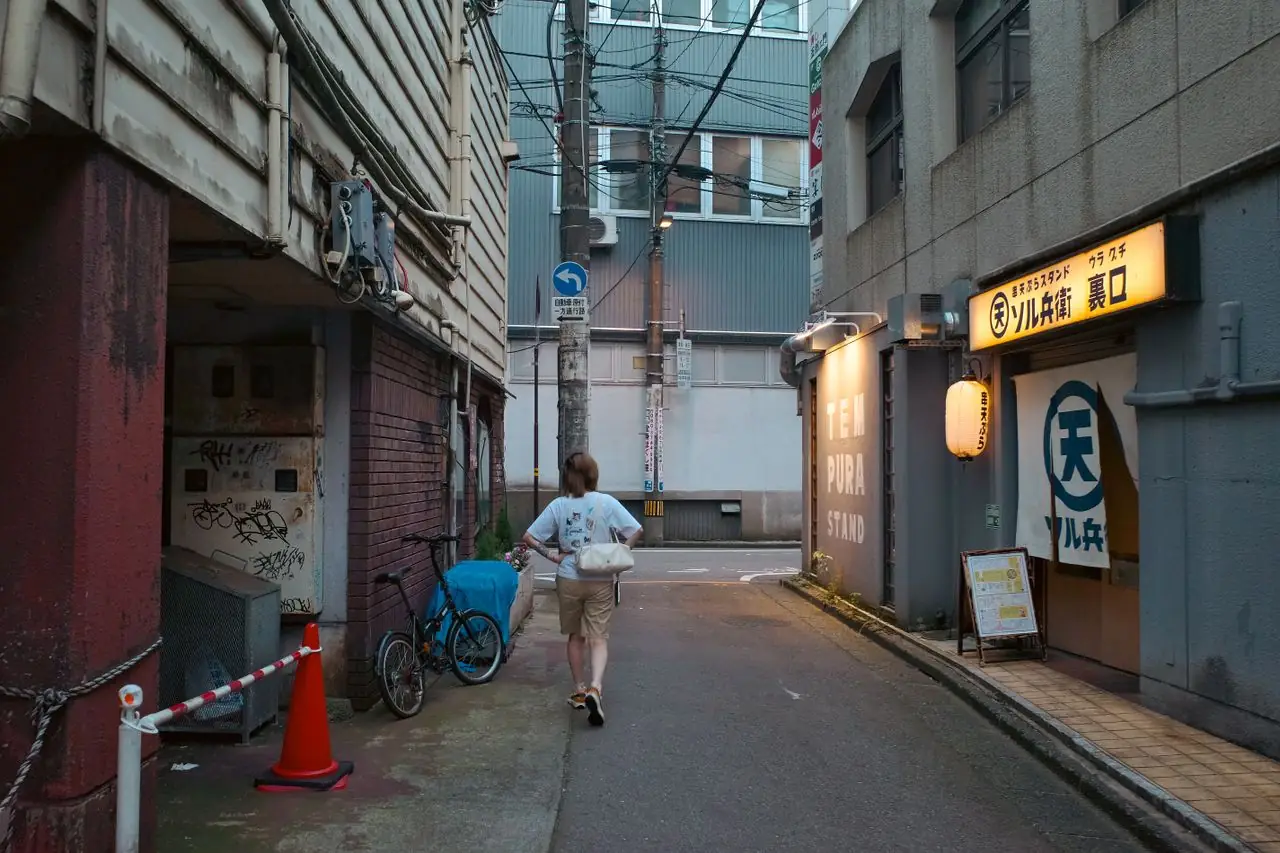 A woman walking alone down a quiet Japanese backstreet past a tempura stand