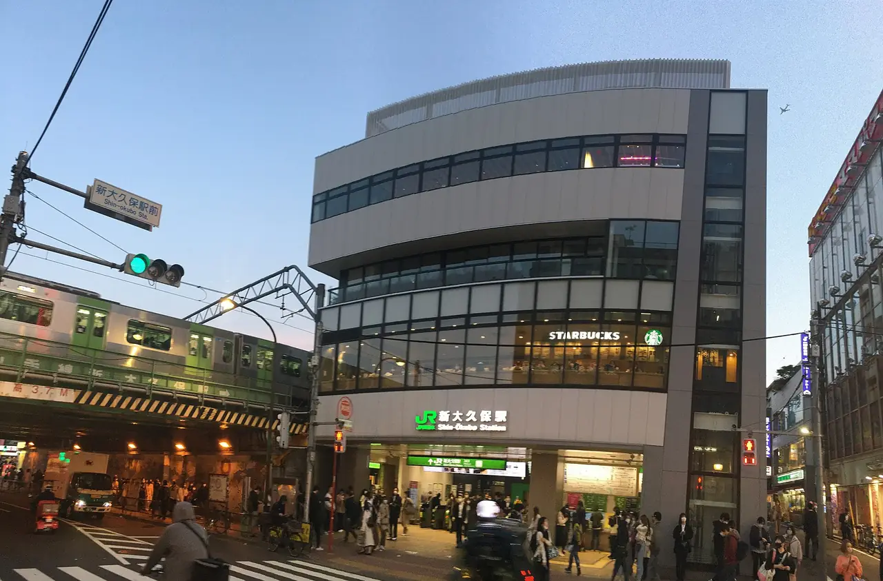 Shin-Okubo Station building with a Yamanote Line train passing at dusk
