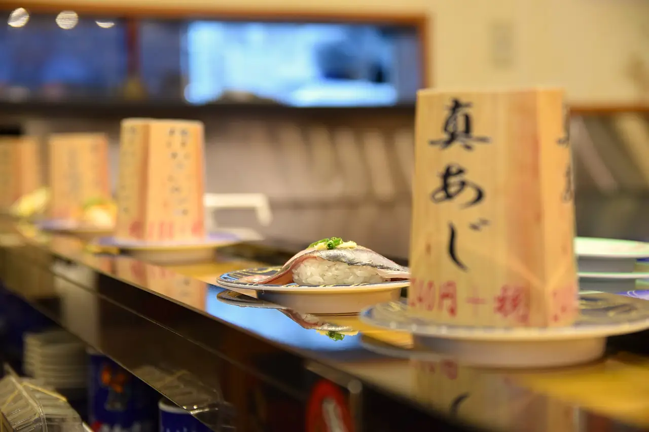 A plate of aji nigiri beside wooden price tags on a kaiten-zushi conveyor belt