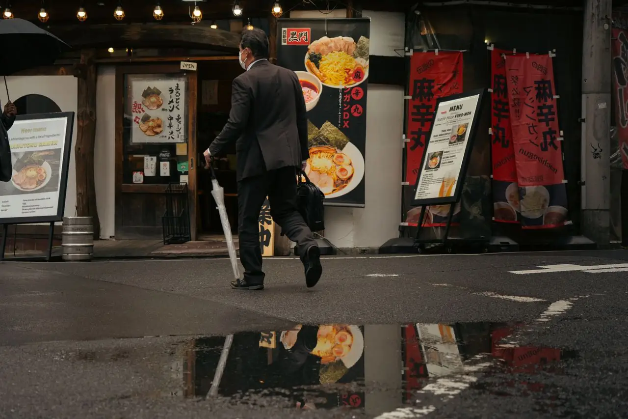 Man with an umbrella walking past a ramen shop entrance on a rainy Tokyo street